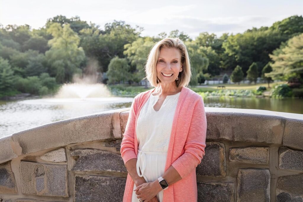 Liz Gabert in a white dress and pink cardigan stands on a stone bridge, smiling at the camera. Behind her is a lake with a fountain. The scene is surrounded by green trees and a partly cloudy sky.